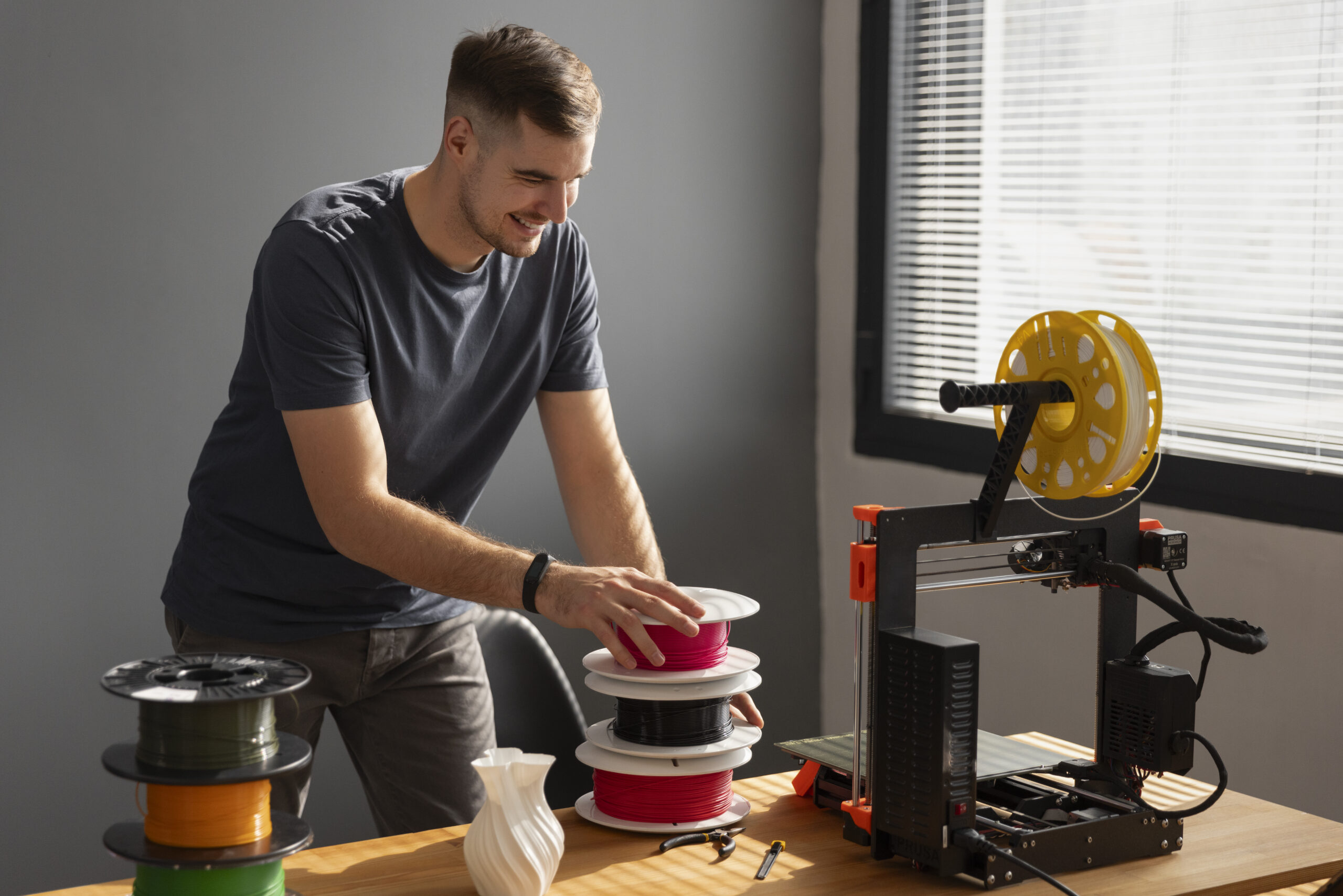 Homme utilisant une imprimante 3D FDM avec différents types de filaments sur un bureau d’atelier.
